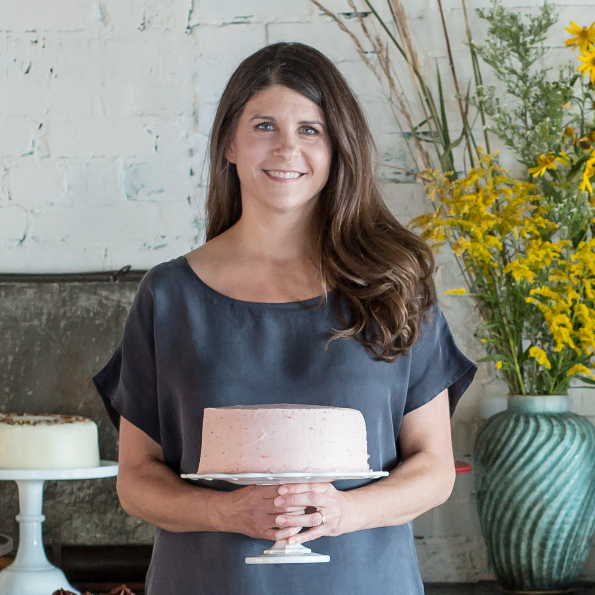 Mary Jennifer Russell, with long brown hair, holds a Sugaree’s Strawberry Cake with pink frosting on a white stand in a studio setting with a white brick wall, dried flowers, and a green vase of yellow flowers