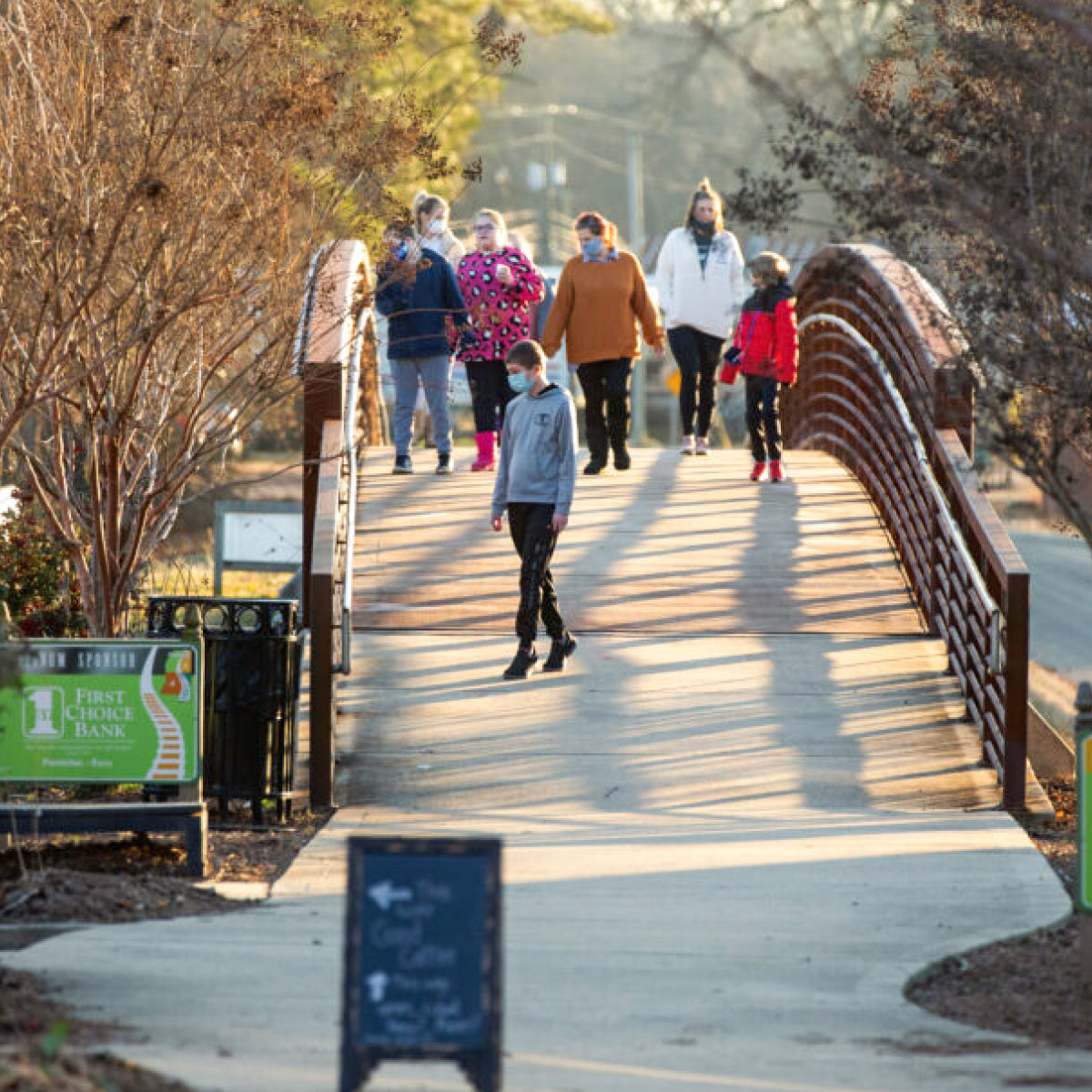 A group of people walk across a small arched wooden bridge on the Tanglefoot Trail in New Albany, Mississippi during late afternoon sunlight, with trees and a sidewalk visible.