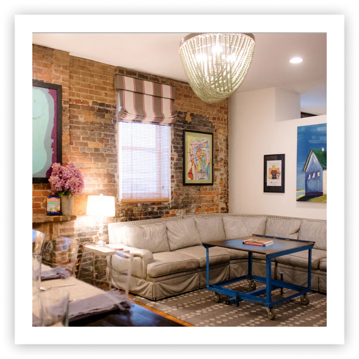 The cozy living room of Sugaree’s downtown loft getaway featuring a light beige sectional sofa, a blue metal coffee table, a brick accent wall, framed artwork, and a large hanging chandelier