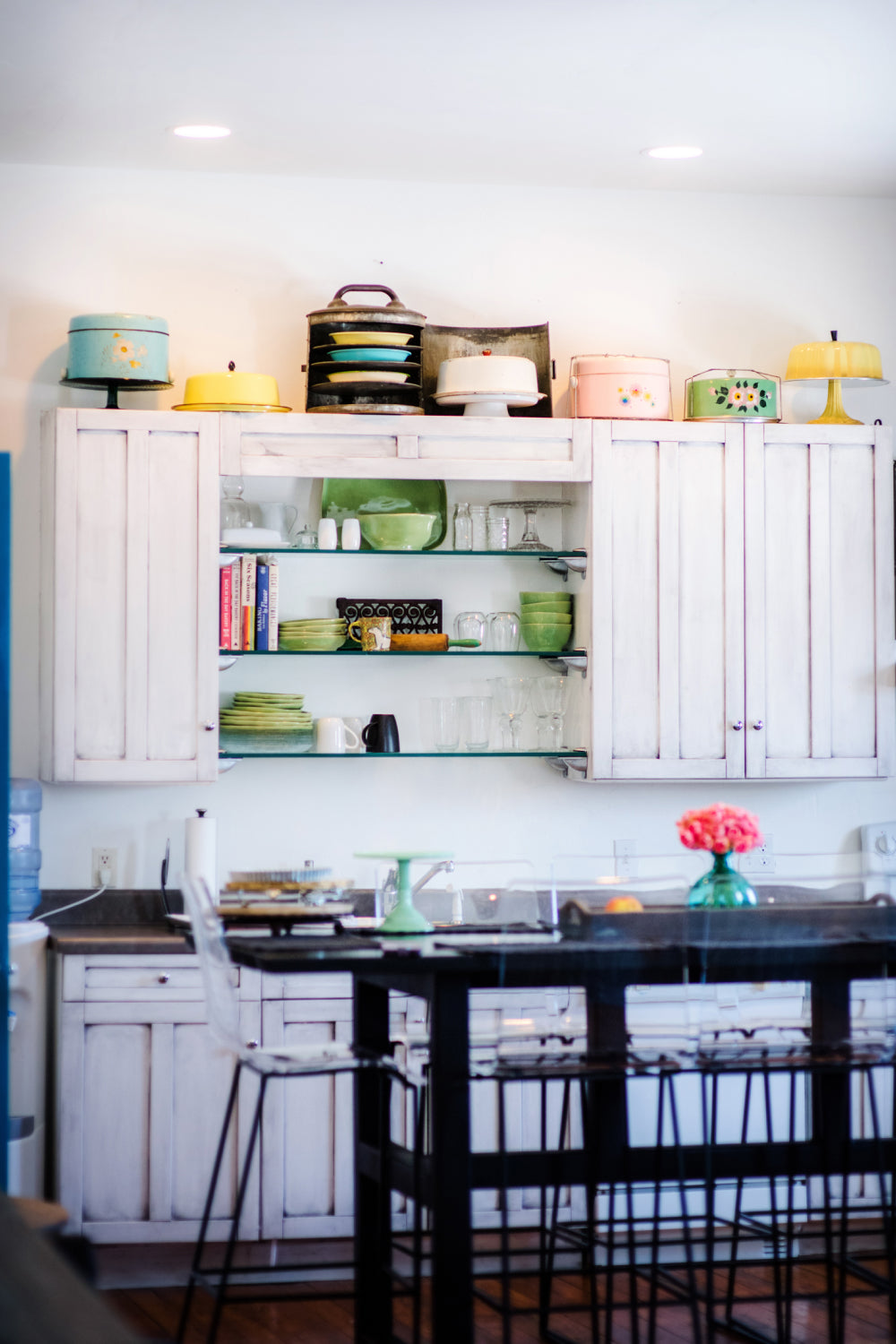 The bright kitchen interior of Sugaree’s downtown loft with white shaker-style cabinets, open shelving with glass shelves, and colorful dishes and decorative items on top. A black dining table with metal bar stools is in the foreground, and a vase of pink flowers sits on the table.