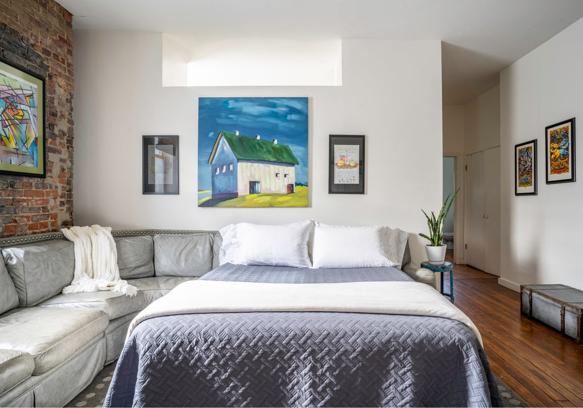 A modern styled sleeping space inside Sugaree’s downtown loft rental features a pullout bed with white and gray bedding, a gray sectional sofa, a brick accent wall, and multiple framed paintings on the walls.
