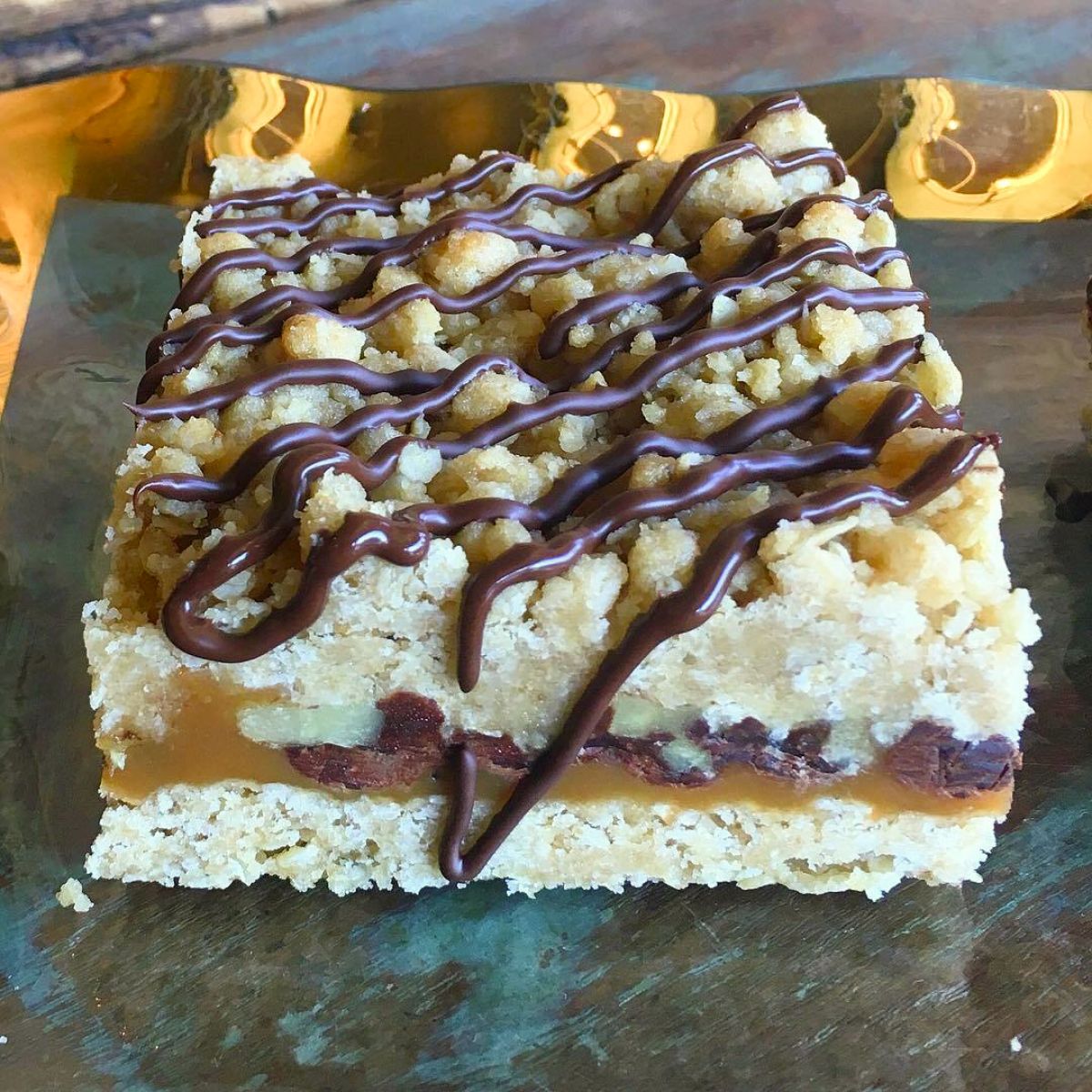 A square sliced Sugaree’s Caramelita bar on a dark surface. The dessert has a golden-brown crumb topping drizzled with dark chocolate, a caramel middle layer, and a light oat base layer with visible walnut pieces.