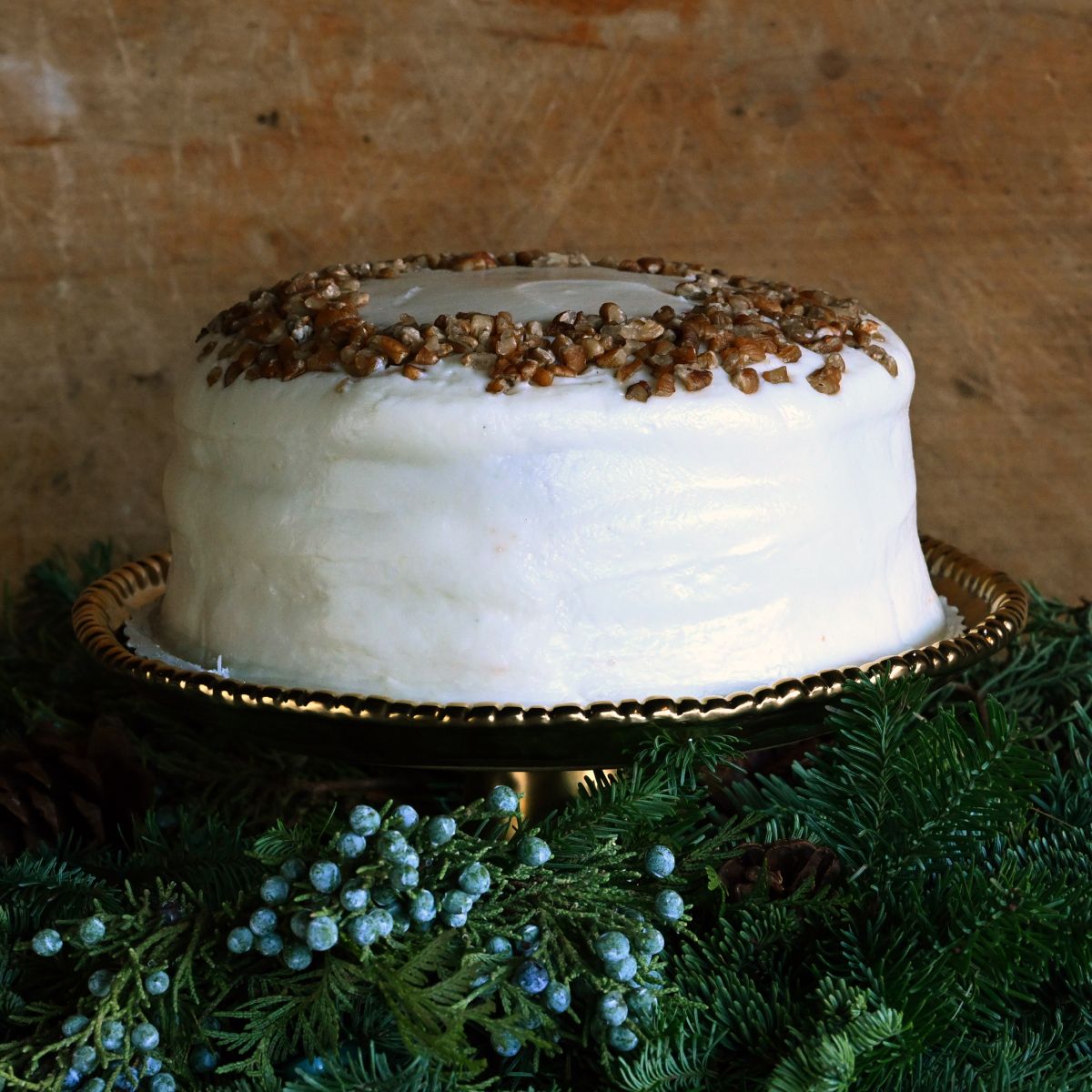 Sugaree’s Italian Cream Cake frosted white with a crumbly pecan topping on a decorative stand with berries and greenery