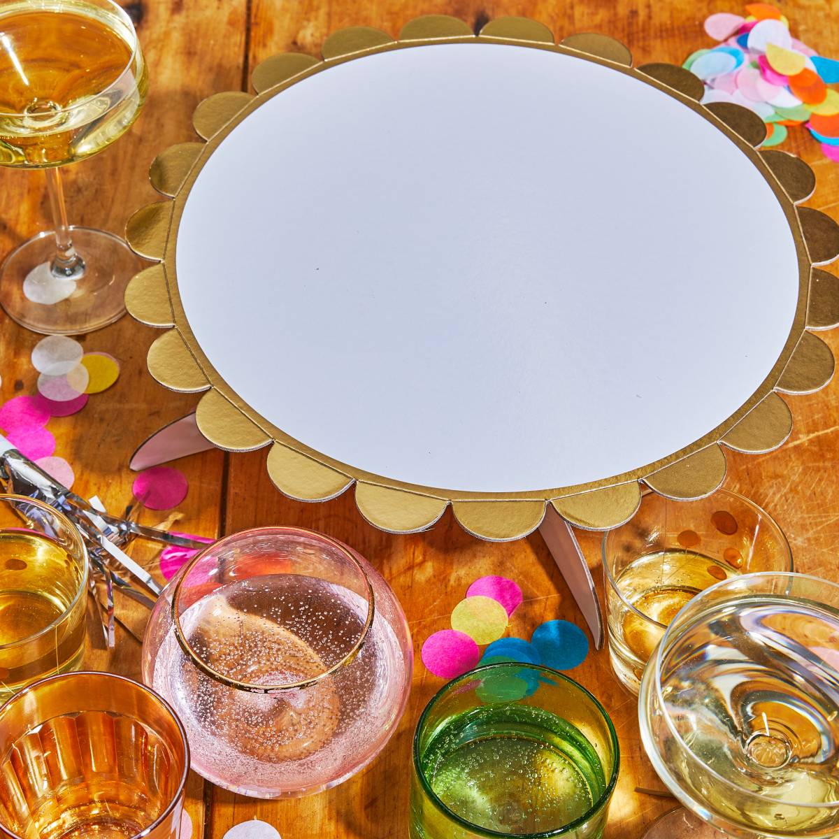 A white cake stand with a scalloped, metallic gold border, styled on a wooden table with drinking glasses, champagne, and colorful confetti