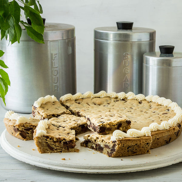 Large chocolate chip cookie on a tray next to stainless steel containers of flour and sugar