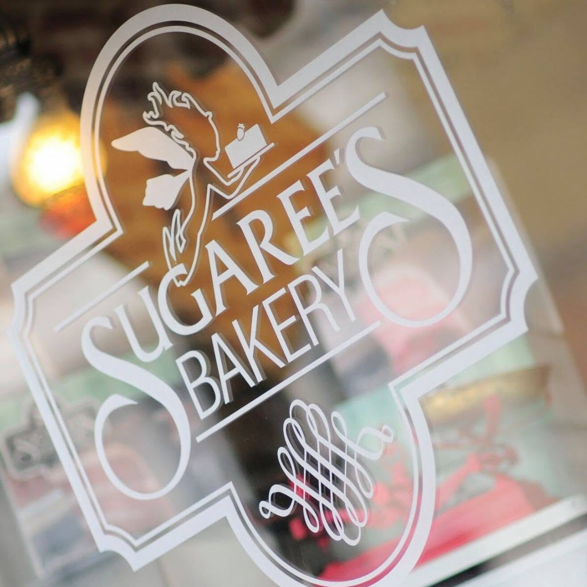 A close-up of a white bakery sign or window decal reading “SUGAREE’S BAKERY” with decorative script and pastry illustrations