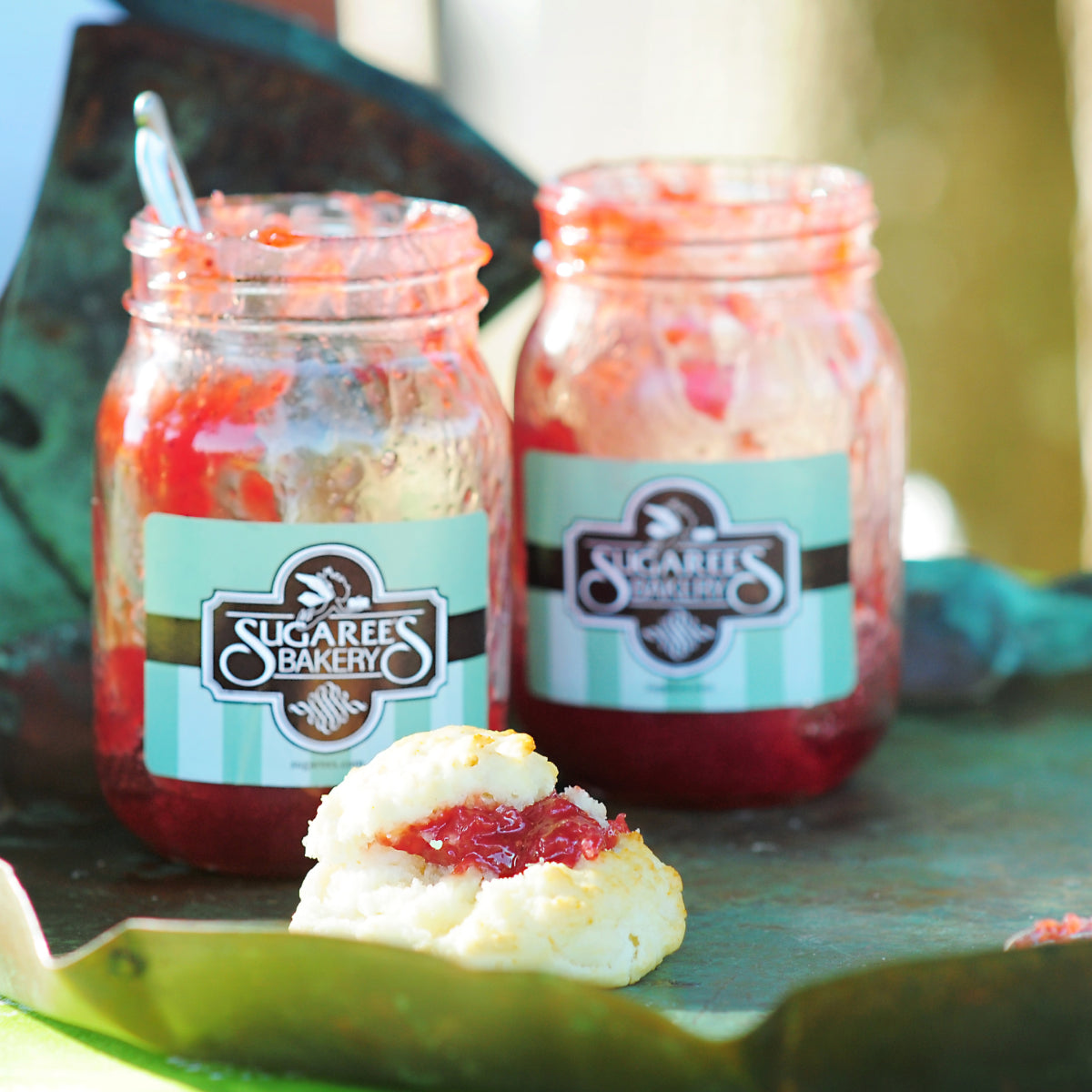 Two glass jars of Sugaree’s Bakery jam sit with a small biscuit in the foreground on a dark surface
