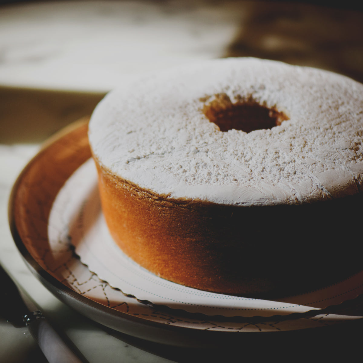 A ring-shaped Sugaree’s Cream Cheese Pound Cake dusted with powdered sugar sits on a decorative plate with a white paper liner in a softly lit setting.