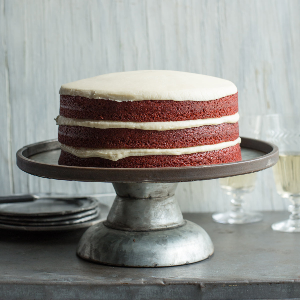 A three-layer Sugaree’s Red Velvet Cake with white frosting sits on a rustic metal cake stand on a dark tabletop with plates and wine glasses blurred in the background.