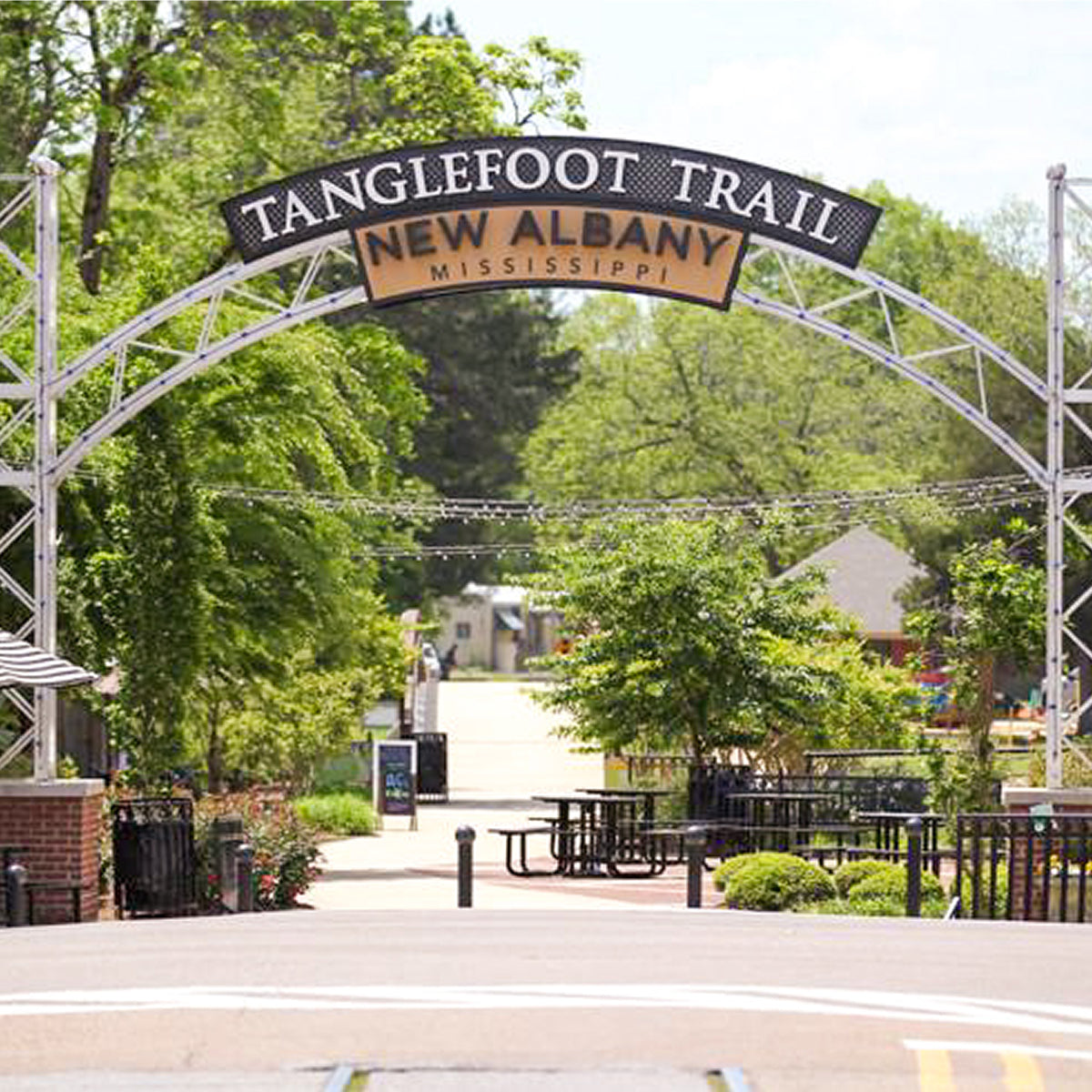 An arched metal sign reads ”TANGLEFOOT TRAIL NEW ALBANY MISSISSIPPI” over a paved path near Sugaree’s Bakery surrounded by green trees and outdoor seating.