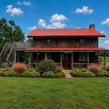 Wooden cabin with a red roof for sale in New Albany, Mississippi. Pictured in a grassy area with flowers and trees under a blue sky.