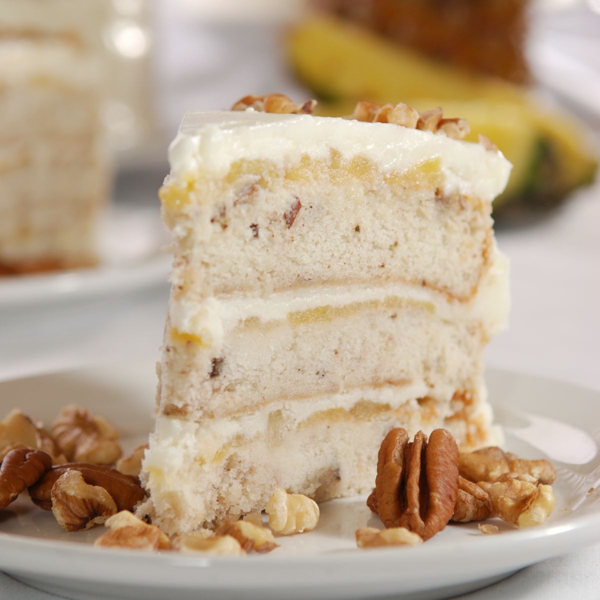 Slice of Sugaree’s Italian Cream Cake showing three layers with white frosting, pecans, and pineapple in background