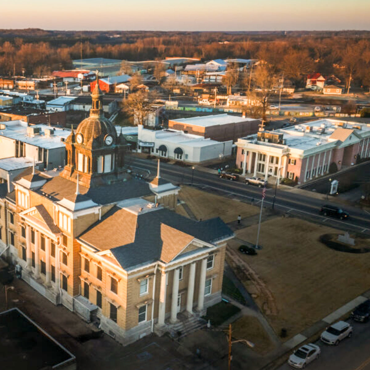 An aerial view of historic downtown New Albany, Mississippi. A building with a central dome and columns is situated among roads, buildings, and trees under warm golden hour light.
