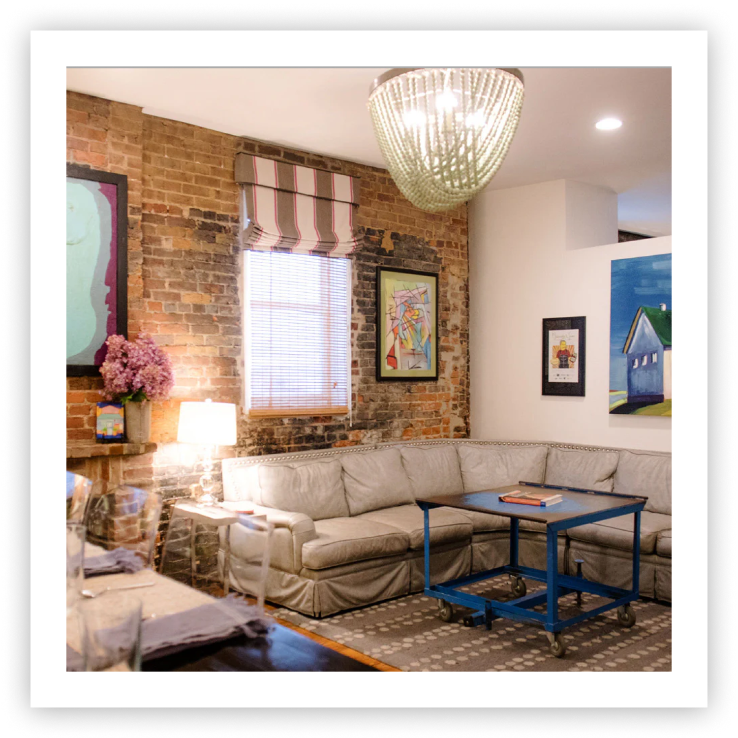 The cozy living room of Sugaree’s downtown loft getaway featuring a light beige sectional sofa, a blue metal coffee table, a brick accent wall, framed artwork, and a large hanging chandelier