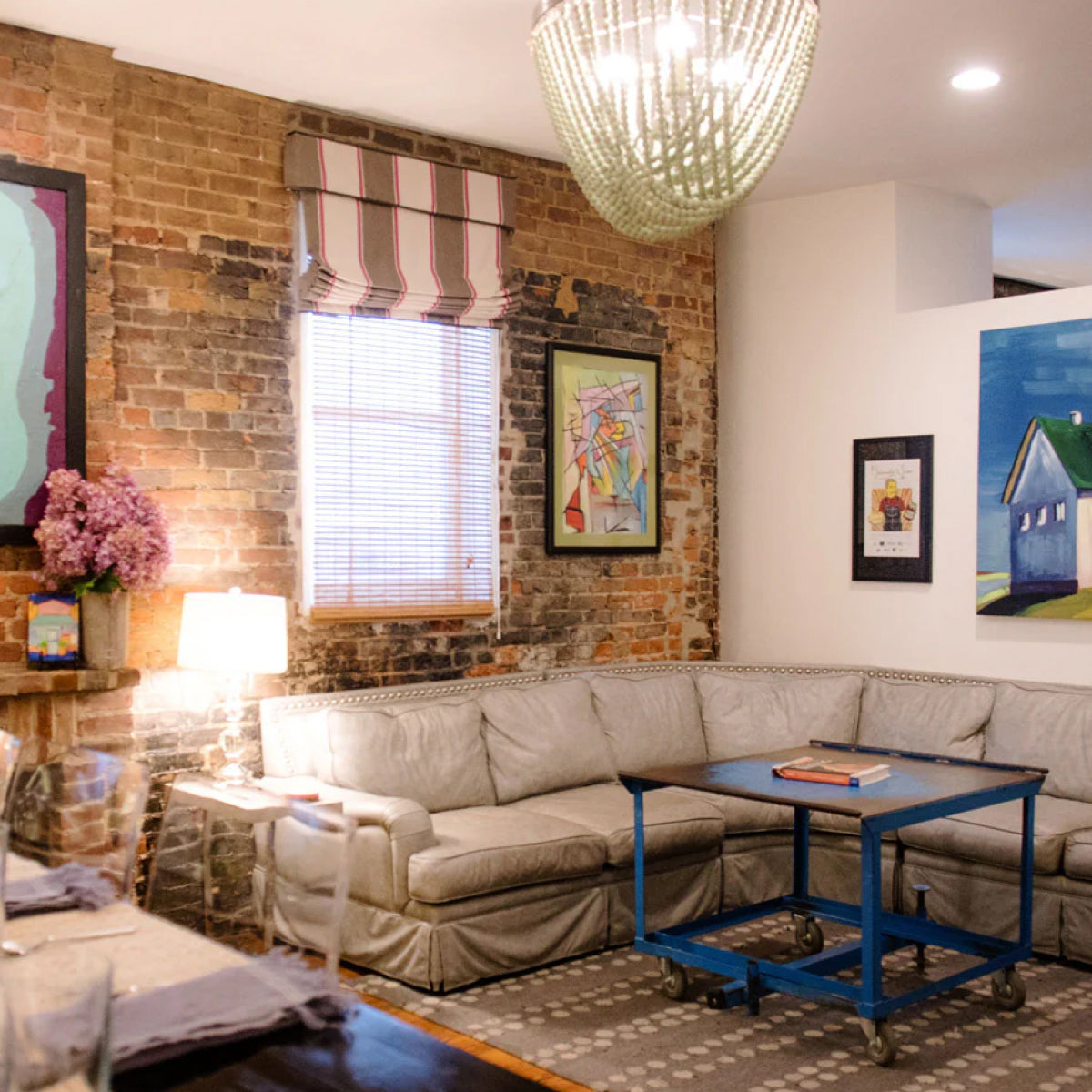 The cozy living room of Sugaree’s downtown loft featuring a light beige sectional sofa, a rustic brick wall, a blue rolling coffee table, and multiple framed art prints on the walls