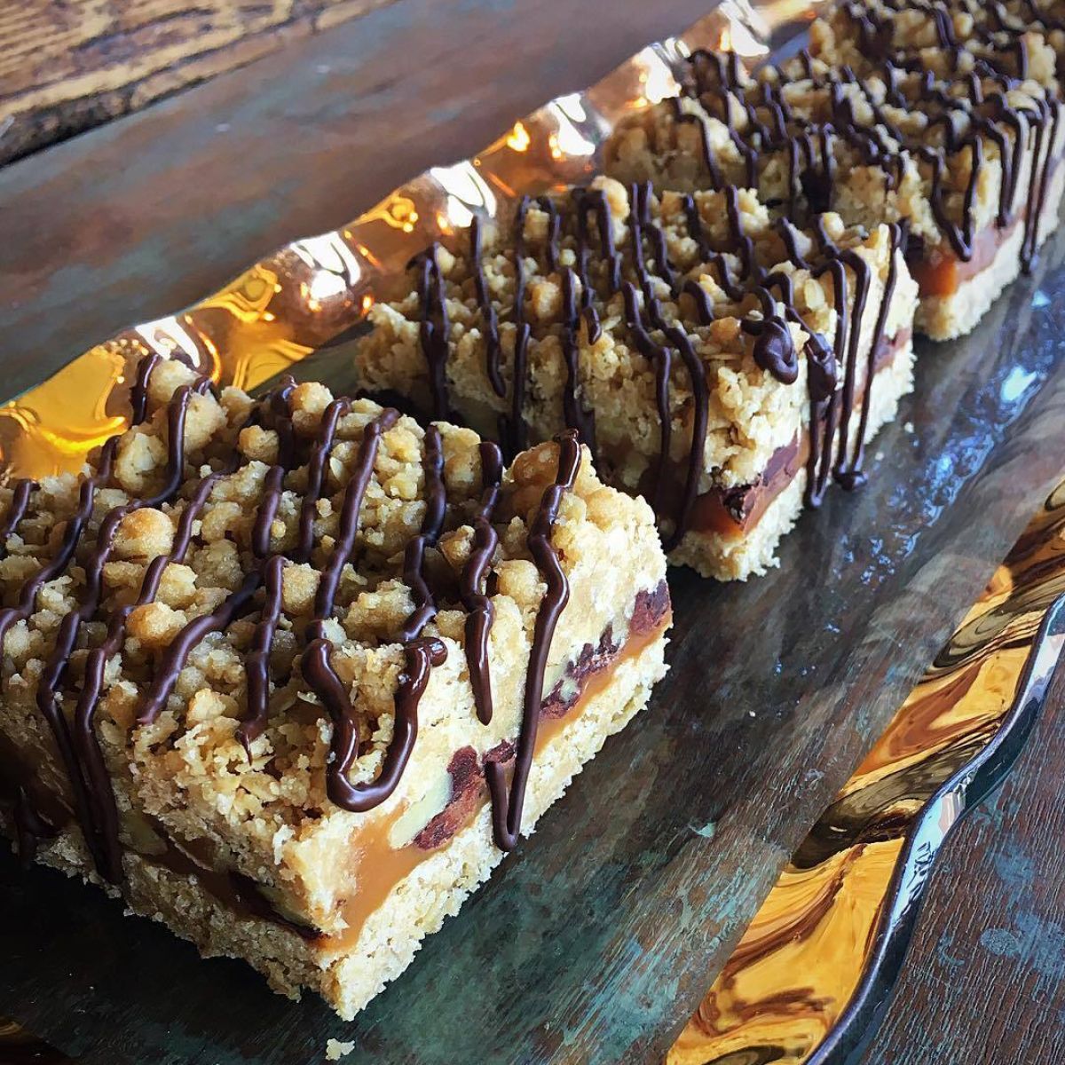 A row of Sugaree’s Caramelita bars topped with crumbles and drizzled chocolate, arranged on a dark wooden board with a gold liner.