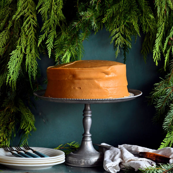 Sugaree’s Caramel Cake on a silver cake stand with greenery in the background