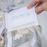 Hand holding a card with 'Sugaree's Bakery' on it, gift basket wrapped in cellophane with silver ribbon.