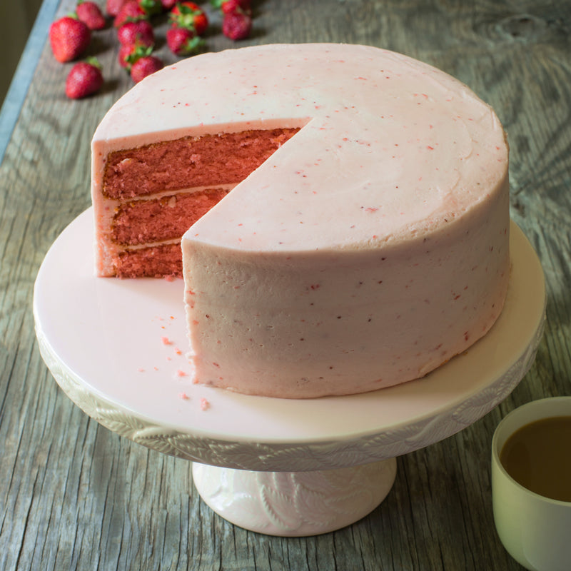 Sugaree’s Strawberry Cake with a slice cut, placed on a white cake stand with strawberries and a cup in the background.