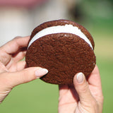 Hands holding a Sugaree’s Homemade Ice Cream Sandwich with chocolate cookies and vanilla ice cream filling, set against a soft, blurred green outdoor background.