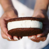 A close-up of a person holding a Sugaree’s Homemade Ice Cream Sandwich with chocolate cookies and vanilla ice cream filling, white shirt visible in the background
