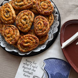 A top-down view of several golden-brown baked Sugaree’s Sausage Pinwheels arranged on a black party tray with a white lace liner. 