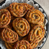 A close-up overhead view of several golden-brown baked Sugaree’s Sausage Pinwheels on a black plate lined with white paper doilies, set on a beige tablecloth